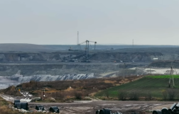 Wielka kopalnia odkrywkowa, z widoczną ogromną koparką wielonaczyniową w centrum, na tle rozległych hałd ziemi i węgla. Na pierwszym planie leżące rury i pola uprawne.