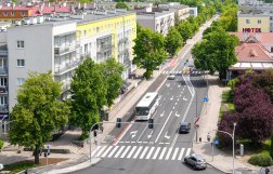 Widok z góry na wielopasmową miejską ulicę z przejściem dla pieszych, po której poruszają się samochody oraz biały autobus. Drogę otaczają zielone drzewa, wielopiętrowe bloki mieszkalne z jasnymi elewacjami oraz budynek z czerwonym dachem i napisem „HOTEL”.