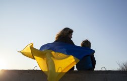 Kobieta i dziecko siedzą tyłem do zdjęcia na murze, okryci są ukraińską flagą.