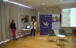 Dwie kobiety podczas prezentacji w sali konferencyjnej. Jedna stoi przy biurku i mówi do mikrofonu, a druga opiera się o ścianę. W tle widoczne są plakaty z logo "Wielkopolska" oraz ekran z wyświetlaną prezentacją.