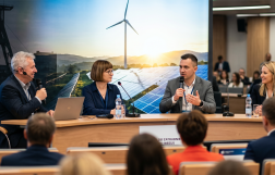 Spotkanie na sali, na zdjęciu widać ludzi na widowni oraz stół prezydialny z siedzącymi przy nim osobami, w tle zdjęcie szybu kopalni i wiatraków