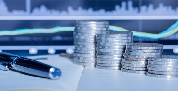 Pióro na otwartym notesie, obok stosy monet, w tle zamazany ekran monitora z wykresem finansowym. Grafika symbolizuje analizę finansową, inwestycje i zarządzanie budżetem.