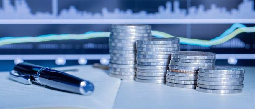Pióro na otwartym notesie, obok stosy monet, w tle zamazany ekran monitora z wykresem finansowym. Grafika symbolizuje analizę finansową, inwestycje i zarządzanie budżetem.