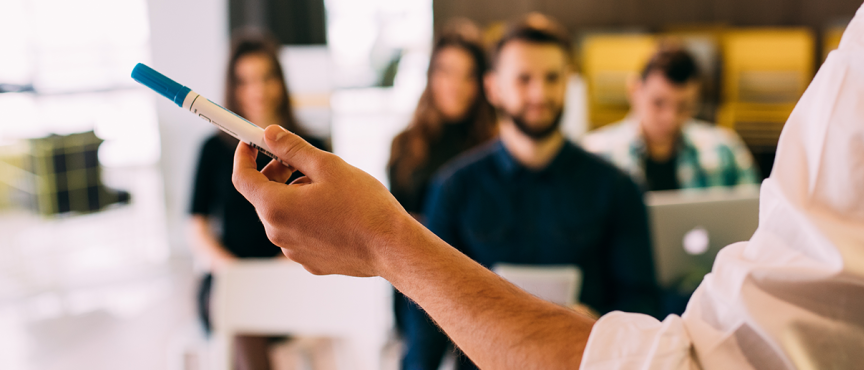 grupa osób siedzących w sali, na pierwszym planie widać uniesioną rękę z długopisem w dłoni