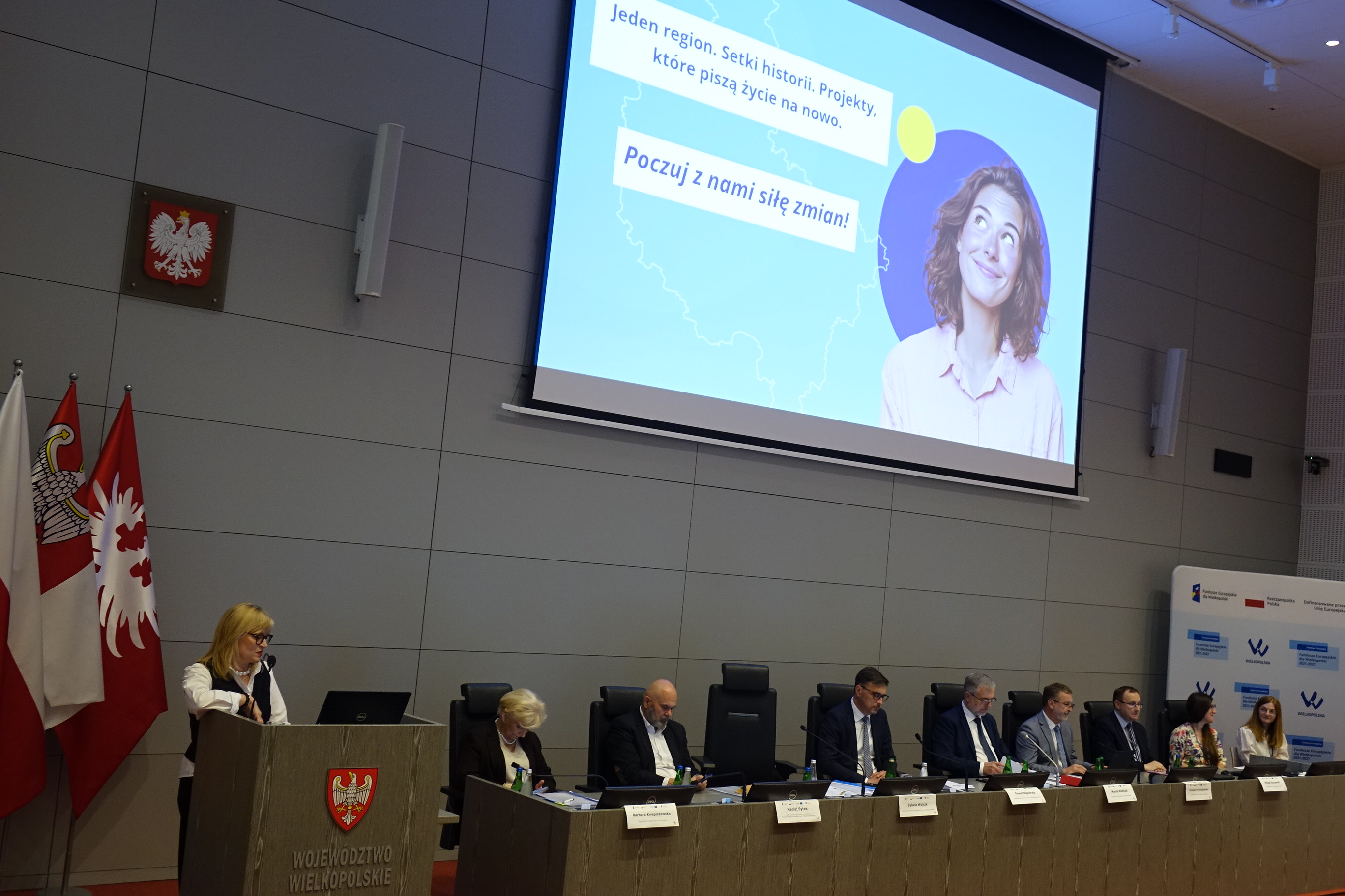 Kobieta przemawiająca w mównicy, na ekranie duży slajd z napisem Poczuj z nami siłę zmian